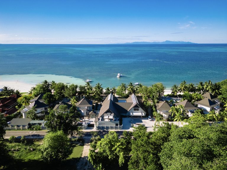Luftaufnahme der Indian Ocean Lodge mit mehreren Gebäuden, Palmen, Straße und Küstenlinie bei Tageslicht