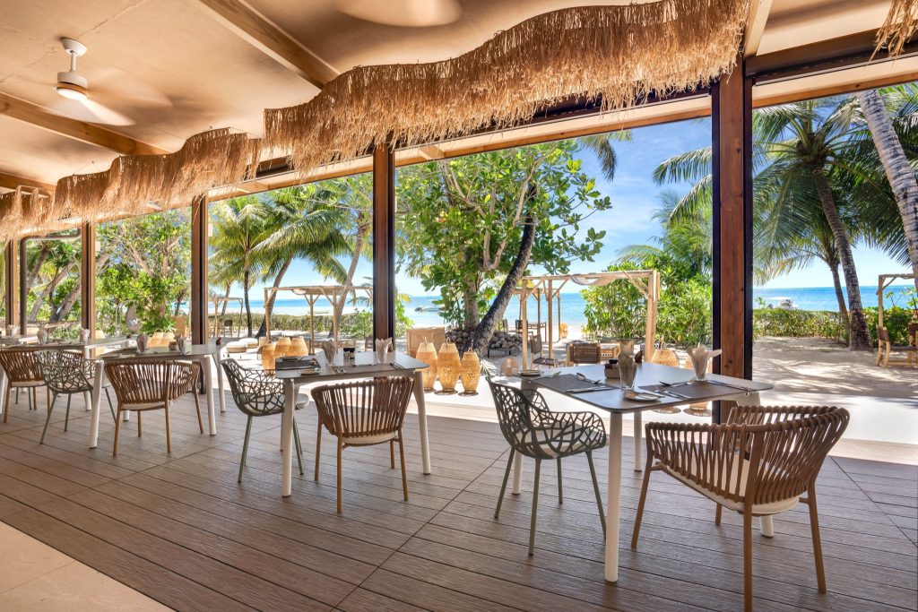 Offenes Restaurant Kokosöl Veranda mit Geflechtstischen, Holzfußboden und Blick auf Strand und Palmen