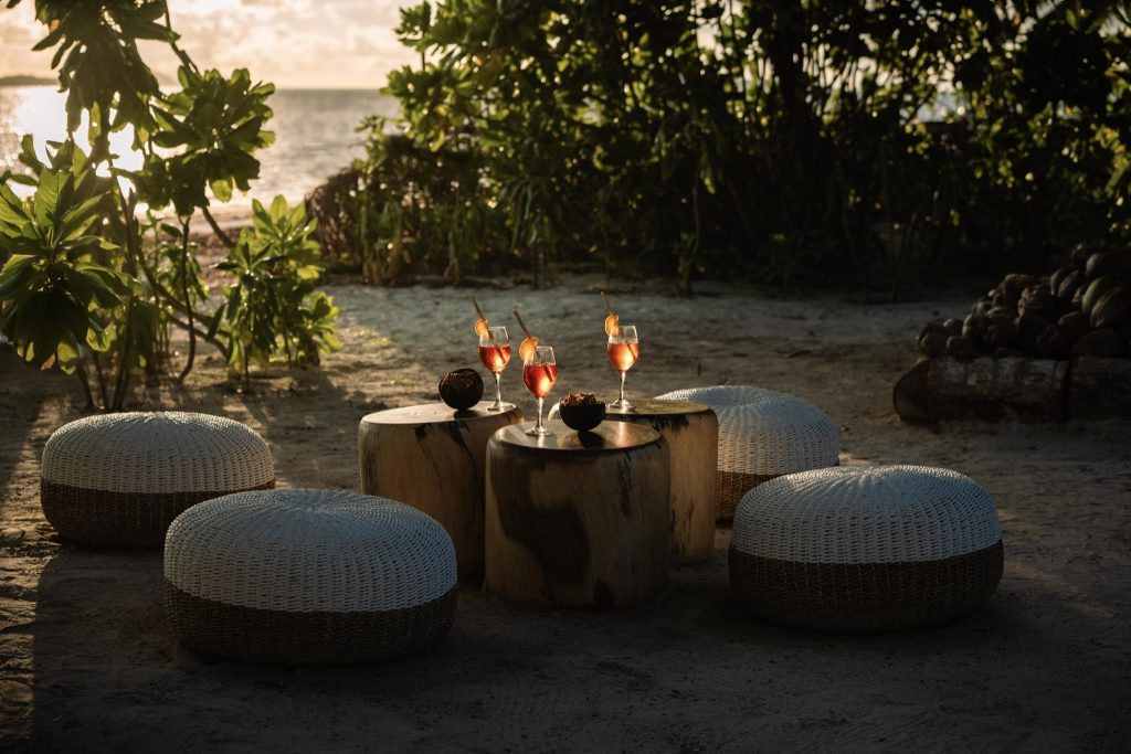 Strand der Indian Ocean Lodge mit runden Holztischen, geflochtenen Sitzkissen und roten Getränken bei Sonnenuntergang