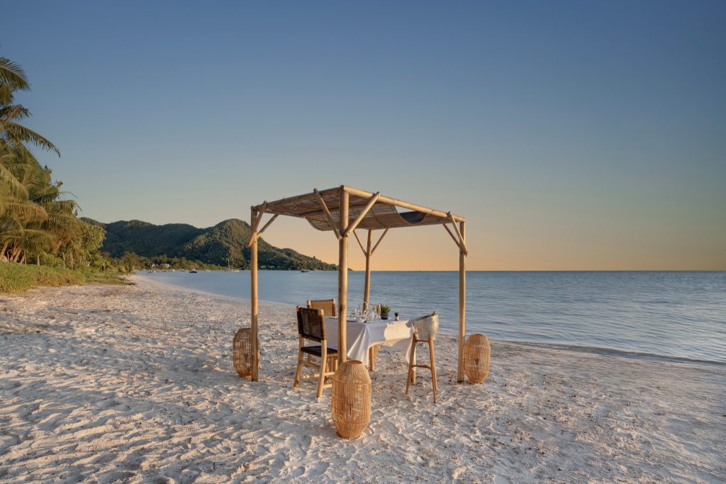 Strand mit Bambuspavillon, gedecktem Tisch und Holzstühlen, grüner Bergkulisse und ruhigem Meer bei Dämmerung