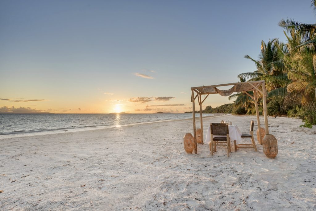 Sandstrand mit Palmen, offene Holzüberdachung und gedecktem Tisch bei Sonnenuntergang an der Indian Ocean Lodge