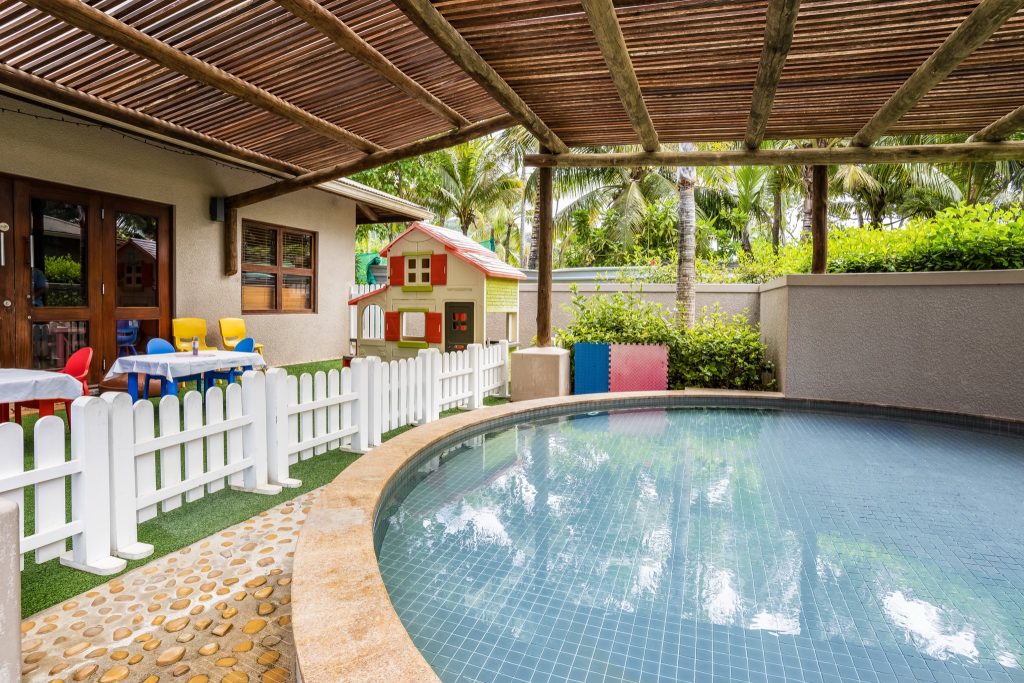 Außenbereich mit kleinem Pool, bunten Kinderstühlen, Spielhäuschen und Palmen im Hintergrund auf den