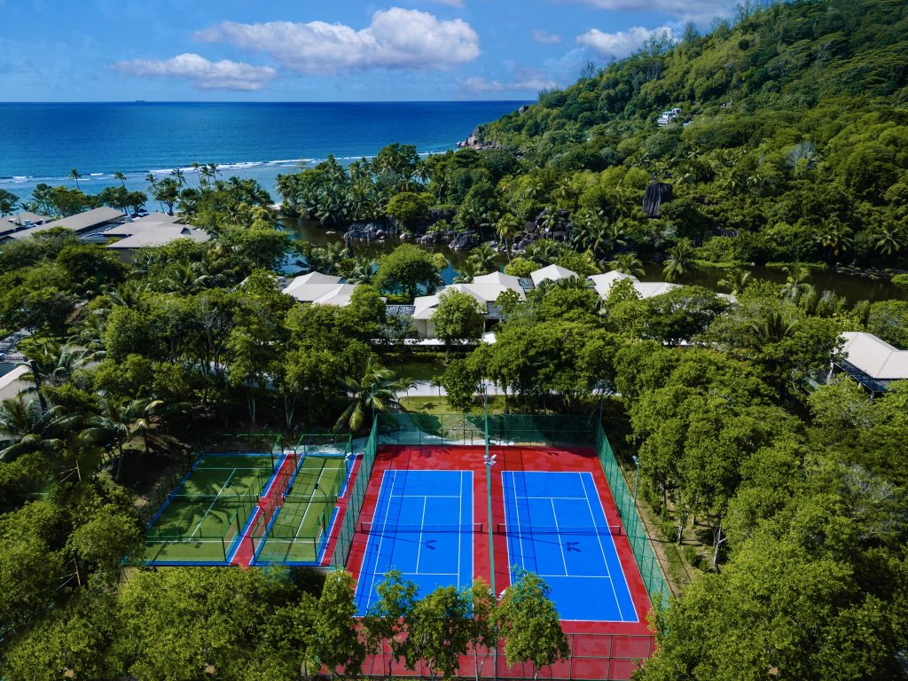 Vogelperspektive auf zwei Tennisplätze mit blau-rotem Belag, umgeben von Vegetation und Meer im Hintergrund