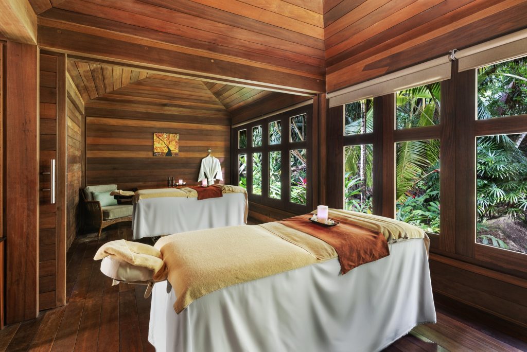 Spa-Raum mit zwei Massagetischen, Kerzen auf Tabletts, Holzdecke und großen Fenstern mit Blick auf grüne Vegetation