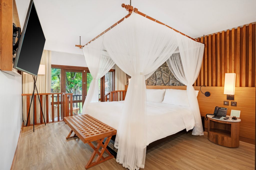 Hotelzimmer Deluxe Lagoon View Room mit Himmelbett, Holzfußboden, Steinwand und großen Fenstern mit Blick ins Grüne