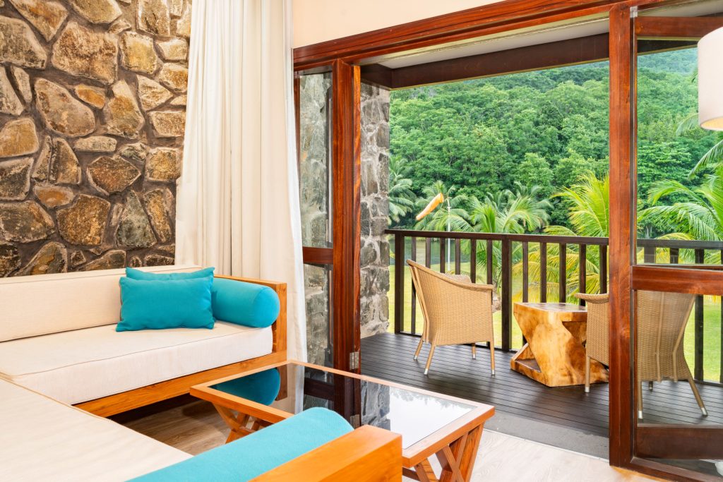 Innenraum eines Deluxe Lagoon View Rooms mit Sofa, Natursteinwand und Balkon mit Rattansesseln, grüne Palmen im Hintergrund