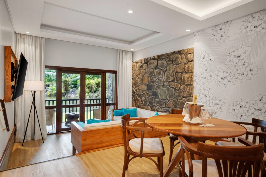 Deluxe Lagoon View Room mit Holz-Esstisch, Sitzbereich mit türkisfarbenen Kissen und großer Glasfront zur tropischen.