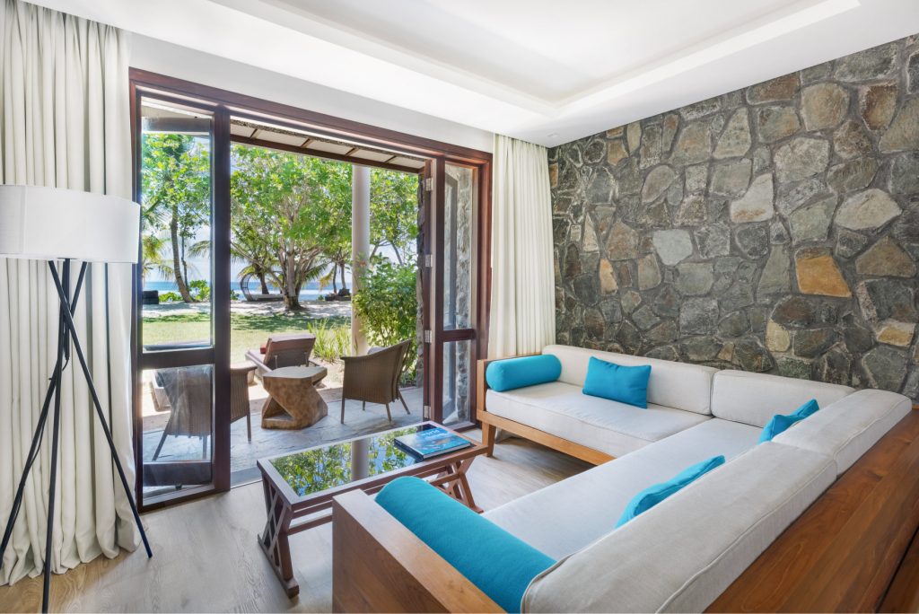 Deluxe Ocean View Garden Room mit Natursteinwänden, L-förmiger Couch und großer Fensterfront im Kempinski Seychelles Resort