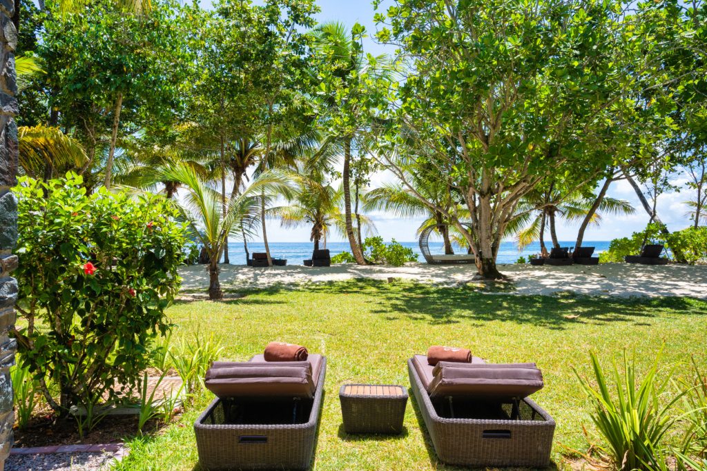 Zwei braune Rattan-Liegestühle mit Handtüchern und Tisch im Garten mit Palmen und Meerblick im Kempinski Seychelles Resort