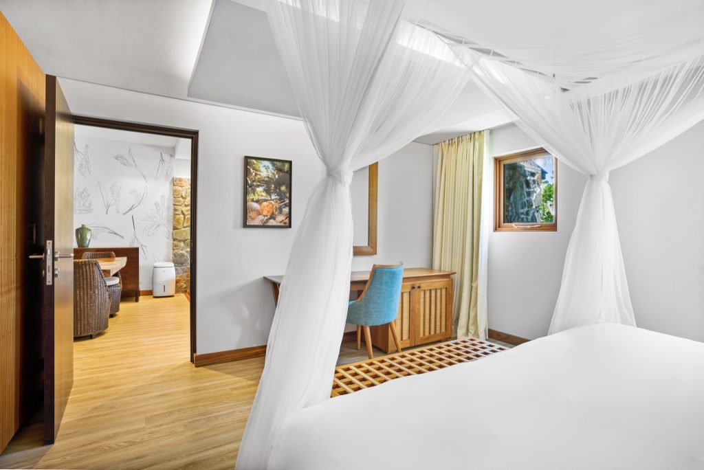 Zimmer der Deluxe Ocean View Suite im Kempinski Seychelles Resort mit weißem Baldachinbett, Holzboden und blauem Stuhl