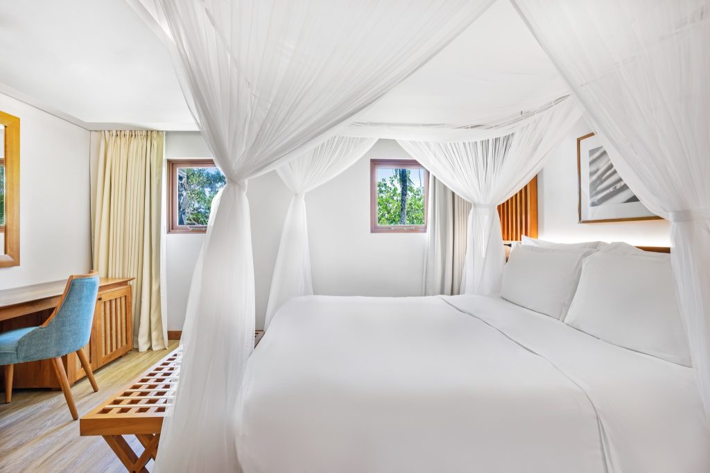 Helles Zimmer mit Himmelbett, weißen Vorhängen, Holzmöbeln und Blick ins Grüne im Kempinski Seychelles Resort