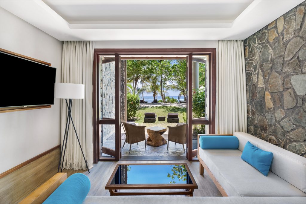 Family Room im Kempinski Seychelles Resort mit hellem Sofa, Natursteinwänden und Meerblick durch große Fenster