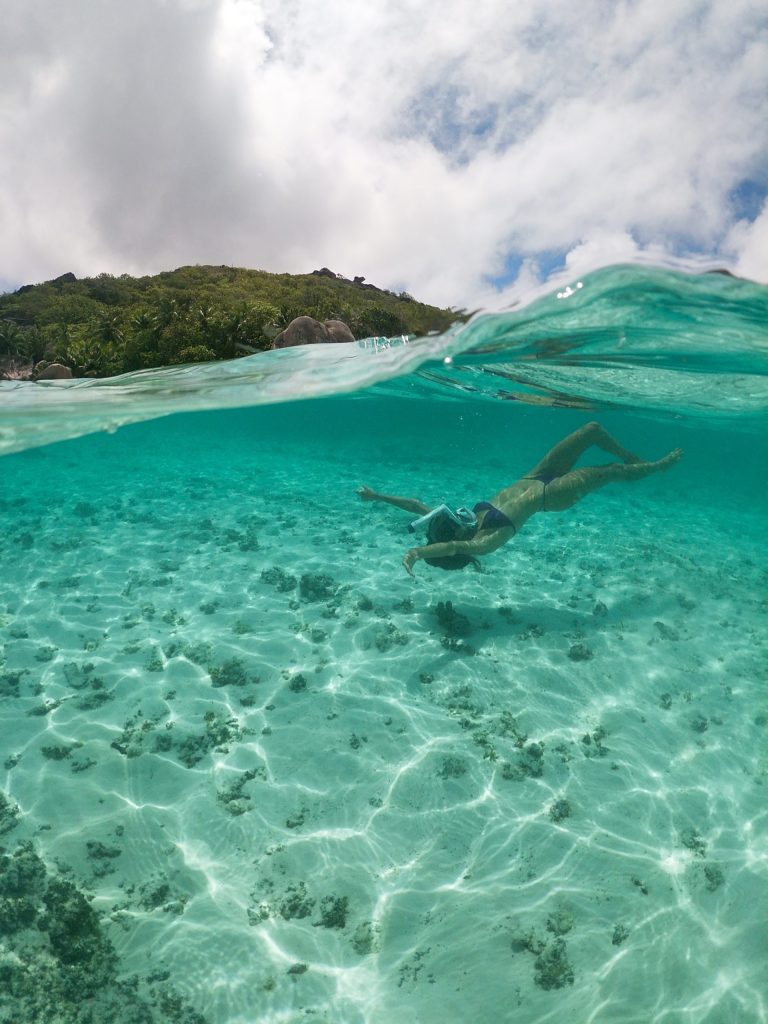 Schwimmer unter Wasser im klaren türkisfarbenen Meer vor grün bewachsenem Hügel bei La Digue Island Lodge