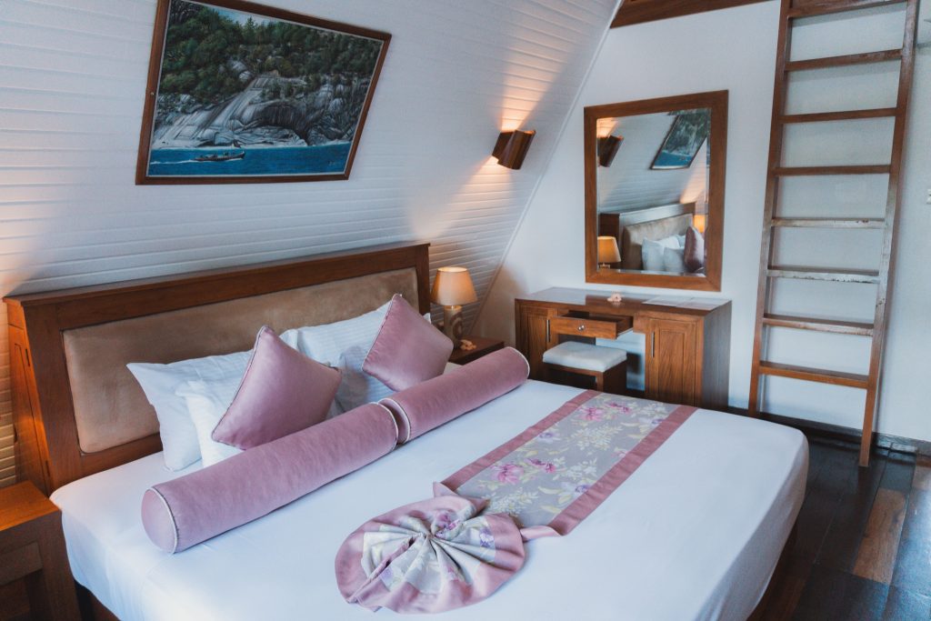 Hotelzimmer Beach Chalet mit großem Bett, weiß-rosa Bettwäsche, Holzkommode, Nachttisch und Wandgemälde mit Meereslandschaft