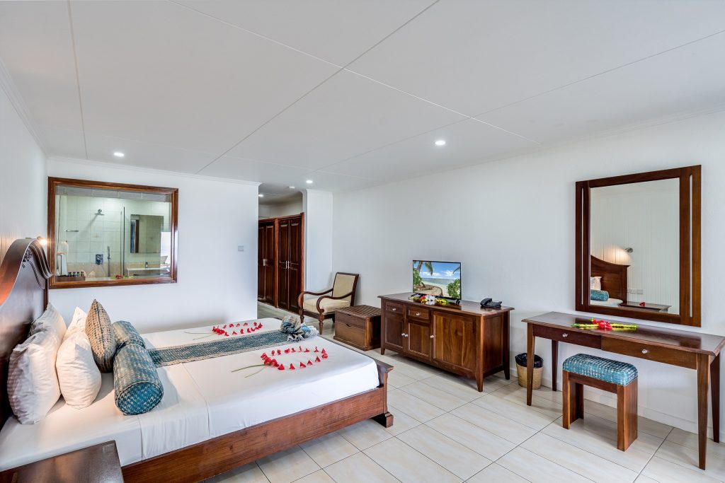Helles Hotelzimmer der Beach House Suite 1BR mit Bett, weißen Laken, blauen Kissen und roten Blütenblättern
