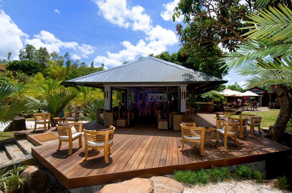 Offene Terrasse der Lakaz Chamarel Exclusive Lodge Bar mit Holzboden, hölzernen Stühlen und dichtem Grün im Hintergrund