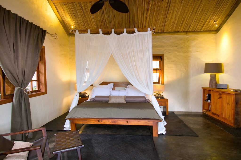 Hotelzimmer mit Holzbett und weißem Baldachin, Holzmöbeln, Beigewänden, Fenster und warmem Licht in Mauritius
