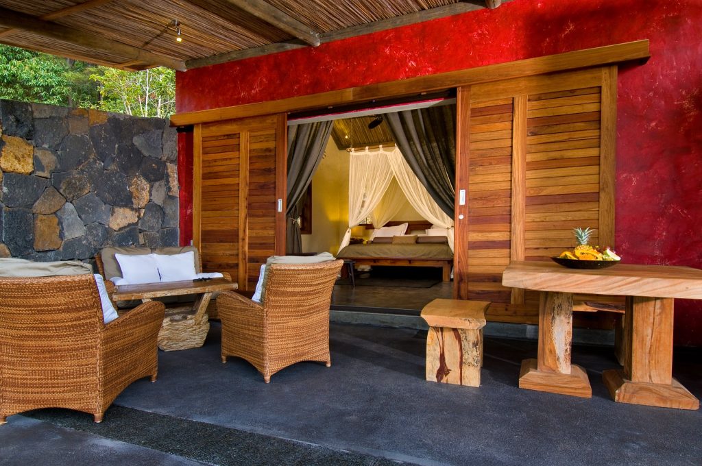 Terrasse mit rustikalen Holzmöbeln und Korbsesseln vor roter Holzplankenwand, Blick ins Zimmer mit Moskitonetz
