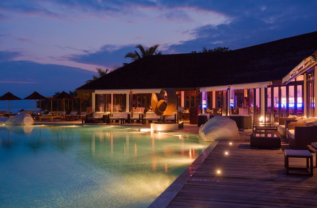 Abendaufnahme der Bar Aqva Pool Bar mit beleuchtetem Holzdeck und Pool im Lily Beach Resort & Spa Malediven