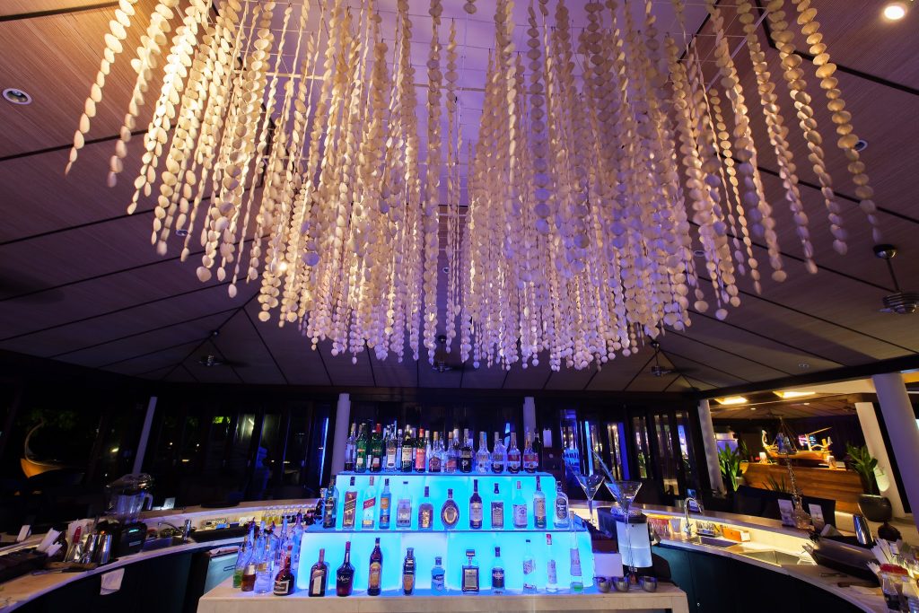 Innenansicht der Spirit Bar im Lily Beach Resort & Spa mit Muschelketten-Deckeninstallation und blau beleuchteten Regalen