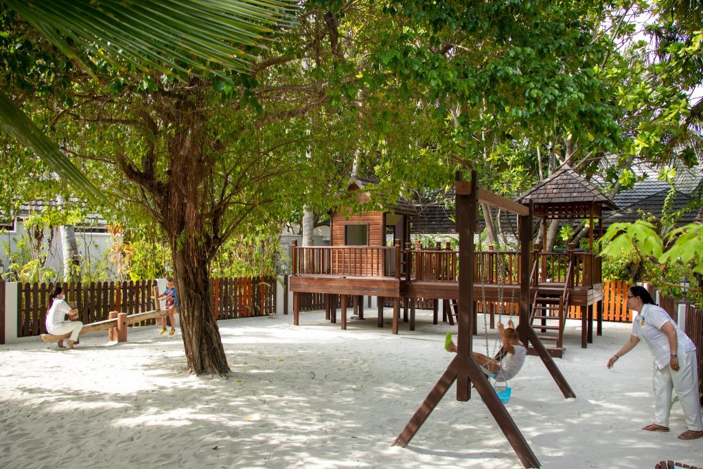 Spielplatz mit großem Baum, Holz-Kletterturm, Sandboden, Schaukel mit Kind und Erwachsenem im Lily Beach Resort