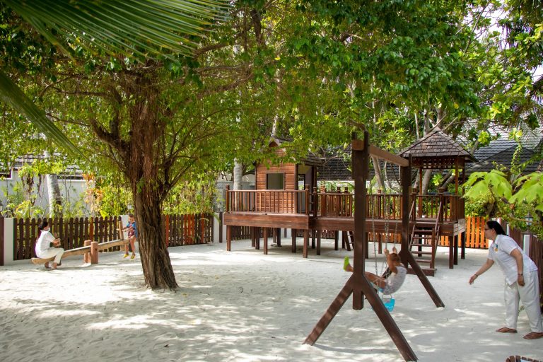 Spielplatz mit großem Baum, Holz-Kletterturm, Sandboden, Schaukel mit Kind und Erwachsenem im Lily Beach Resort