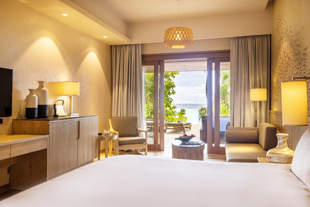 Beach Suite Zimmer mit großem Bett, Anrichte, Tischleuchte, Glasschiebetür und Terrasse im Lily Beach Resort & Spa.