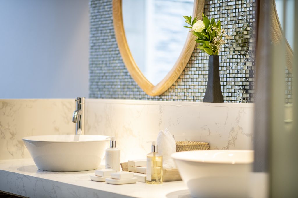 Badezimmer mit zwei freistehenden weißen Keramikwaschbecken, hellem Marmortisch und Mosaikfliesenwand im Lily Beach Resort.
