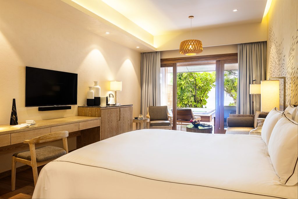 Modernes Hotelzimmer der Beach Suite mit weißer Bettdecke, Schreibtisch, Fernseher und Balkon mit grüner Vegetation