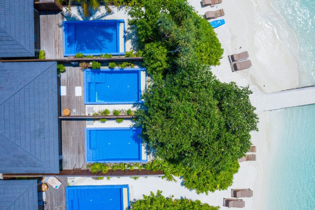 Vogelperspektive auf mehrere rechteckige Swimmingpools mit Holzterrassen und grüner Vegetation am Strand