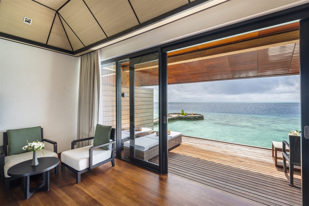 Zimmer Lagoon Villa mit zwei Polstersesseln, rundem Tisch, Holzfußboden und Glasschiebetür zur Terrasse über dem Meer