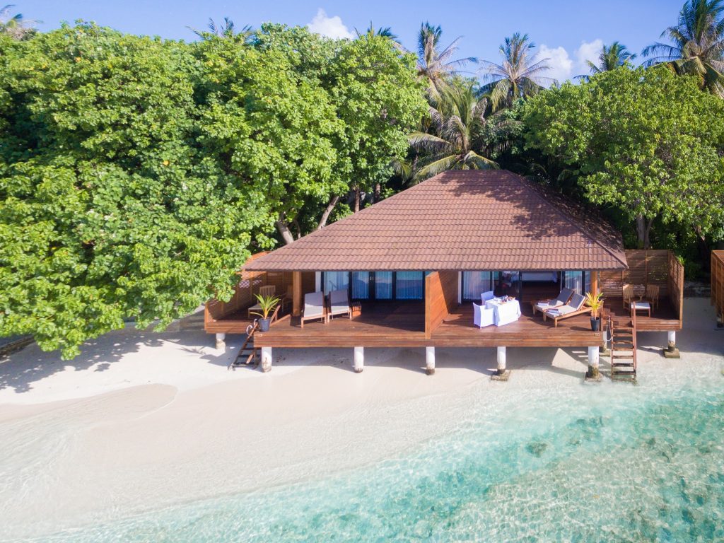 Über Wasser gebauter Holz-Bungalow mit Terrasse und Holzmöbeln, Palmen im Hintergrund auf den