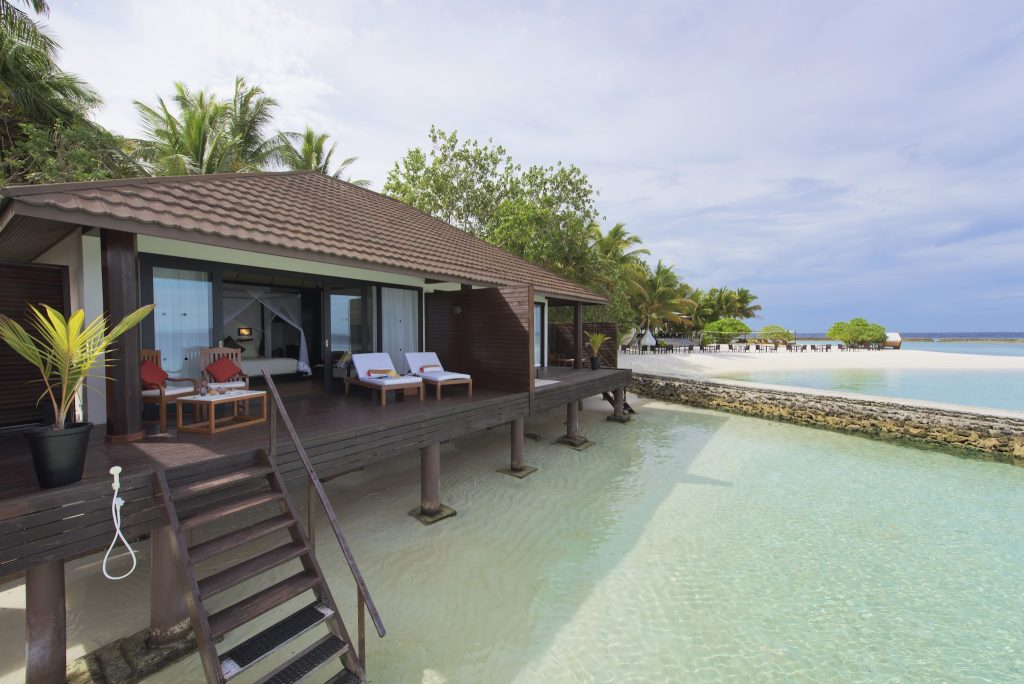 Bungalow auf Stelzen über klarem Wasser mit Holzterrasse, zwei weißen Liegestühlen und gläsernen Schiebetüren
