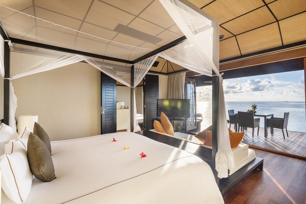 Zimmer der Sunset Water Suite mit Himmelbett, weißen Laken, Holzmöbeln und Terrasse mit Meerblick auf den