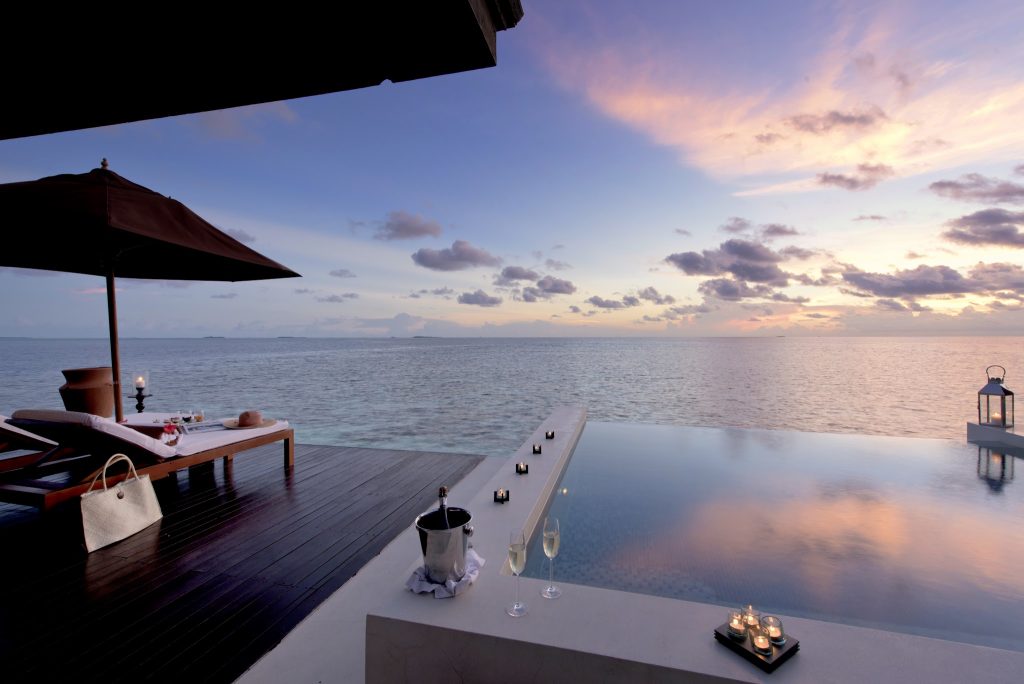 Terrasse an Infinity-Pool mit Holzliege, Sonnenschirm, Kerzen, Sektkühler und Meer im Sonnenuntergang Malediven