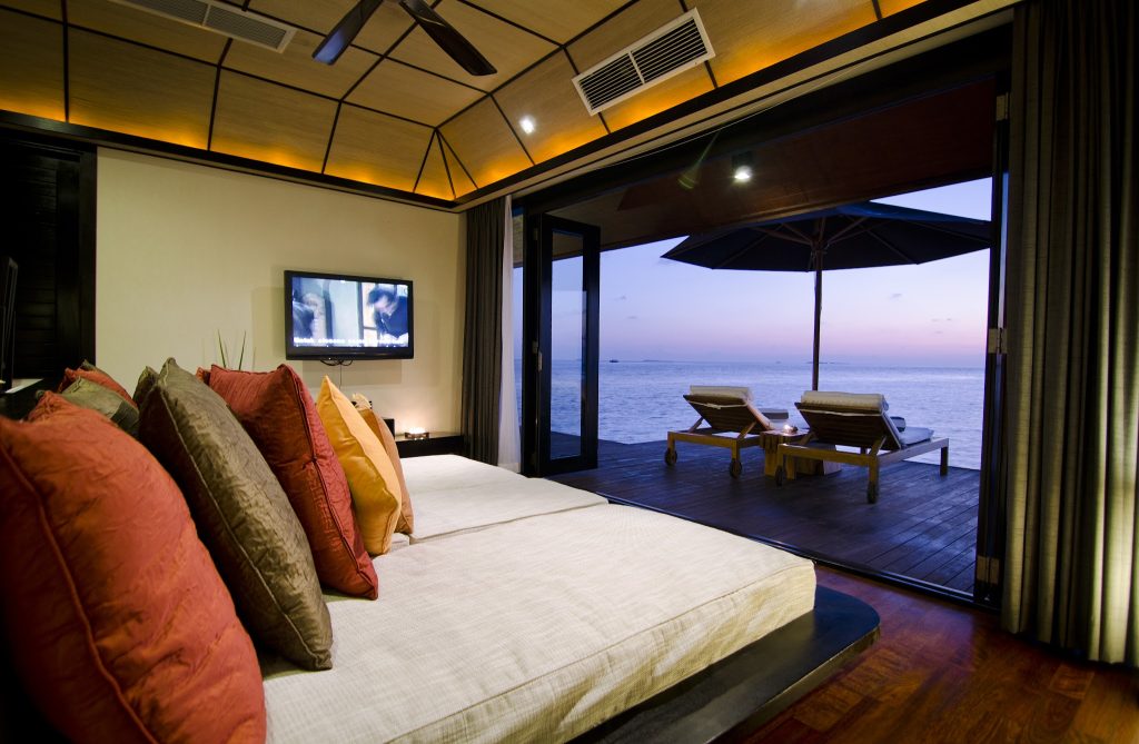 Zimmer der Water Suite mit großem Bett, bunten Kissen und offener Terrasse mit Meerblick bei Sonnenuntergang