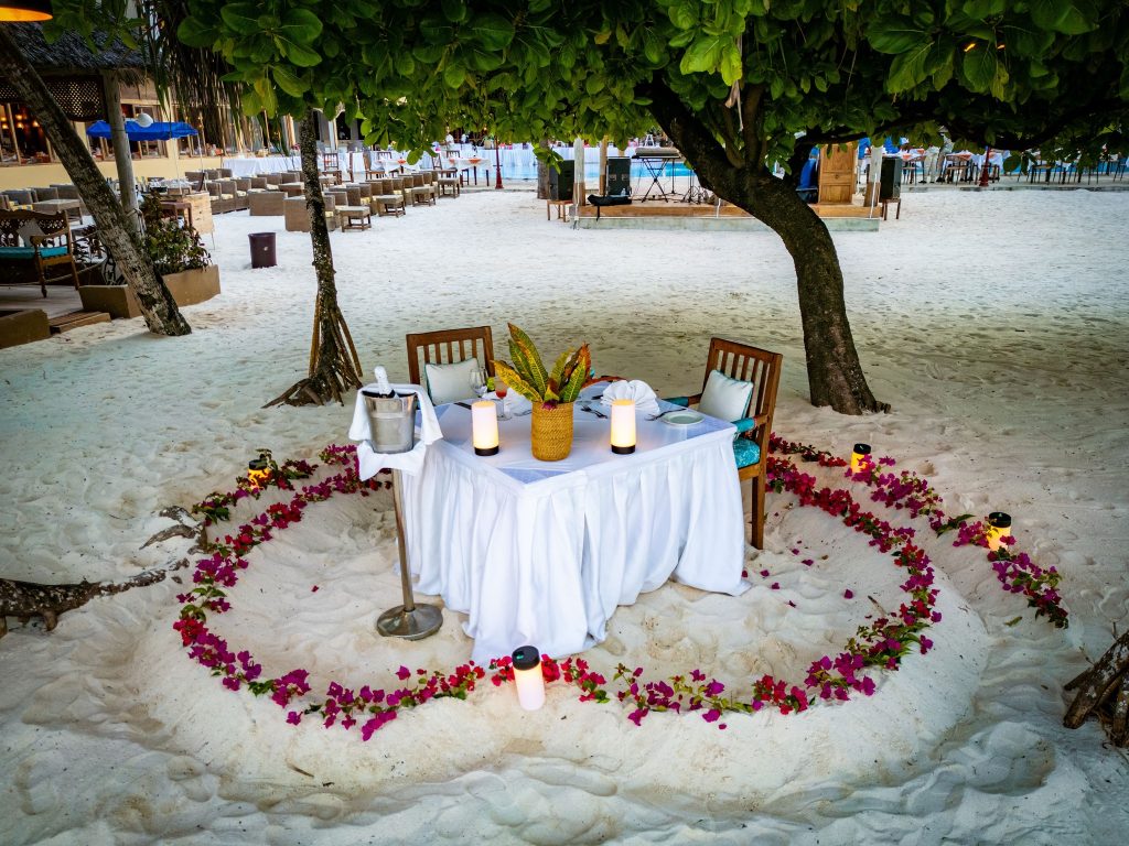 Strand-Dinner-Setup auf weißem Sand mit Tisch, weißen Tischdecken, Kerzen und roten Blütenblättern in Herzform