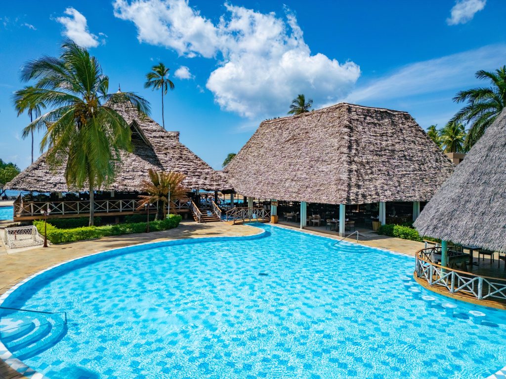 Außenansicht des Neptune Pwani Beach Resort & Spa Pools mit traditionellen Strohdächern und Palmen in Sansibar