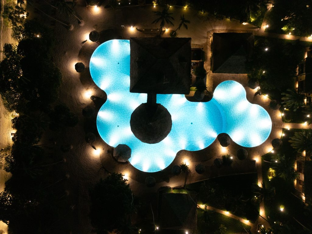 Vogelperspektive auf großen blau beleuchteten Pool mit Strohsonnenschirmen und Wegen bei Nacht