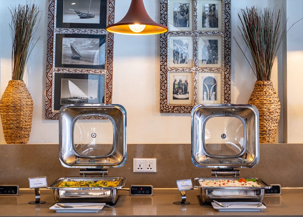 Buffetstation mit zwei offenen Warmhaltebehältern aus Metall, Schildern und dekorativen Vasen im Restaurant Neptune Pwani.