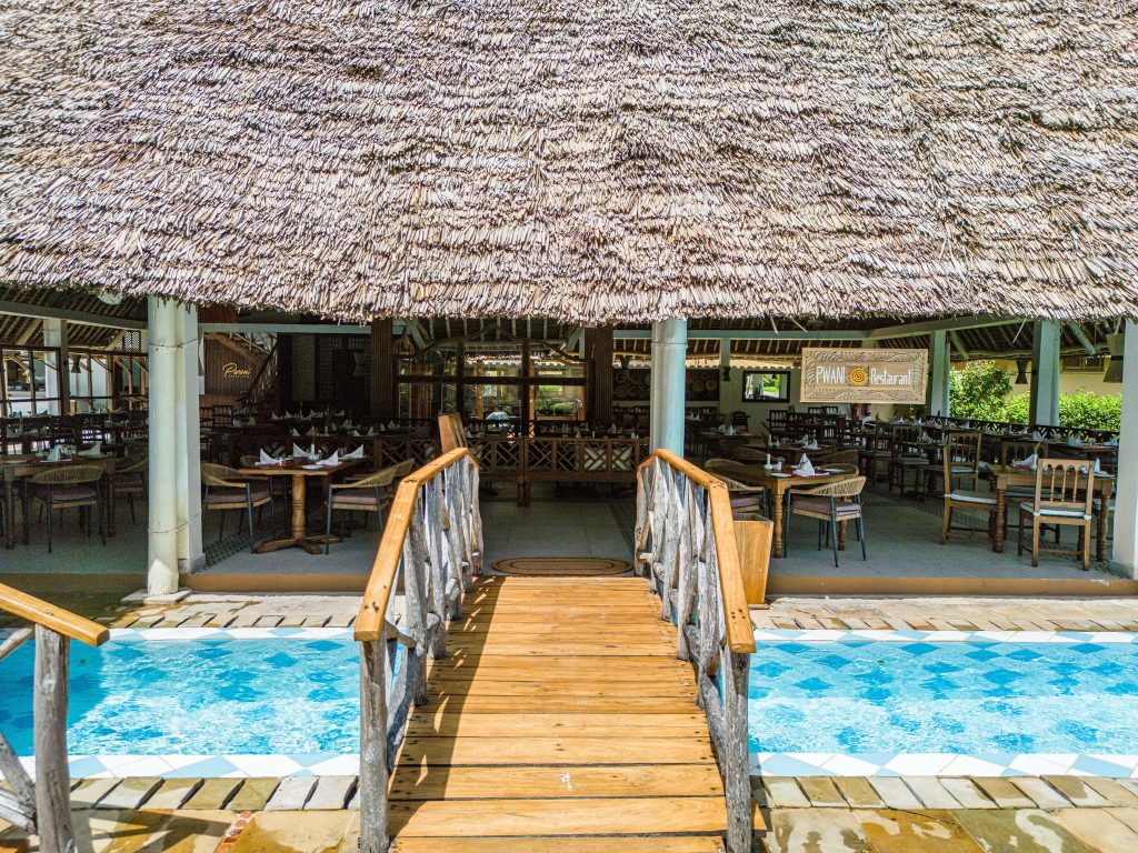 Offener Restaurantbereich im Neptune Pwani Beach Resort & Spa mit Strohdach, Holztischen und hölzerner Brücke über Pool