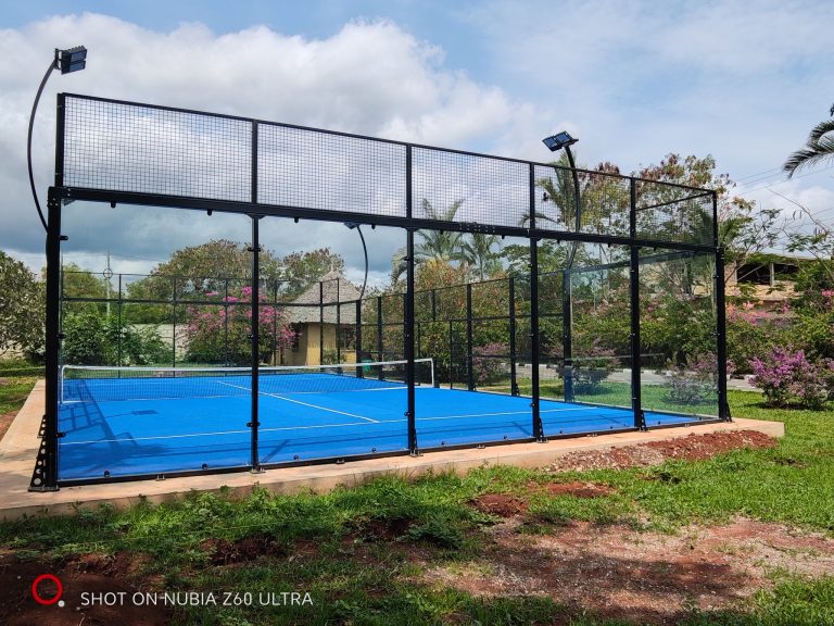 Padel-Platz mit blauem Boden und Metallgitter in Gartenanlage mit Rasen, Sträuchern und strohgedecktem Gebäude