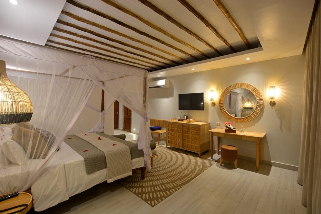 Hotelzimmer mit großem Bett und weißem Moskitonetz, Holzbalkendecke, Kommode, Fernseher und rundem Spiegel im Deluxe Room