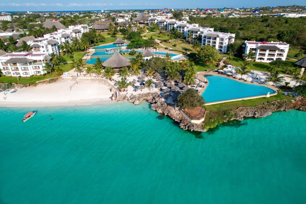 Luftbild des Royal Zanzibar Beach Resort mit weißem Sandstrand, Boot, mehreren Pools, Palmen und mehrstöckigen Gebäuden