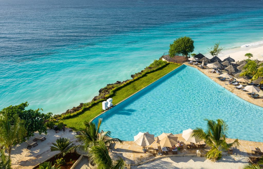 Großer Infinity-Pool mit türkisblauem Ozean im Hintergrund, Liegestühle und Sonnenschirme, Palmen