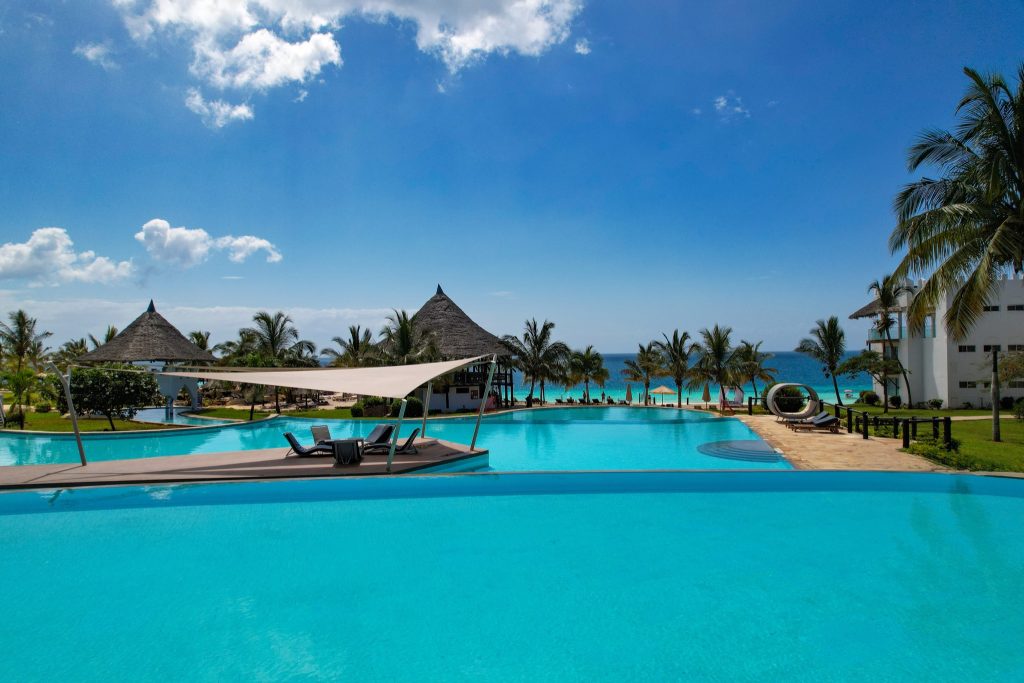 Großer klarer Pool mit modernem Segeltuch-Sonnenschutz und Liegestühlen, Palmen und Strohdachpavillons im Hintergrund