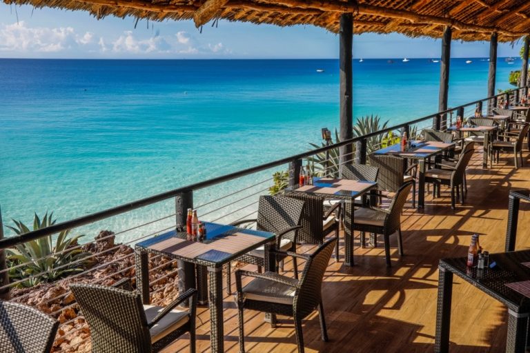 Offene Restaurantterrasse mit dunklen Rattanmöbeln auf Holzboden und Blick auf das blaue Meer in Sansibar