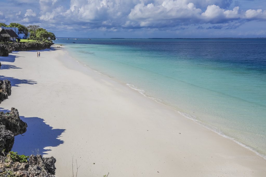 Langer weißer Sandstrand am türkisfarbenen Meer mit Felsen und Strohdachgebäuden, bewölkter Himmel