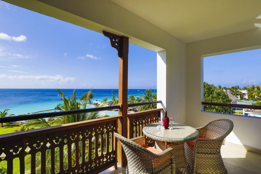 Balkon mit runden Glastisch, zwei Rattanstühlen, rotem Tuch und Weingläsern, Blick auf Meer und Palmen