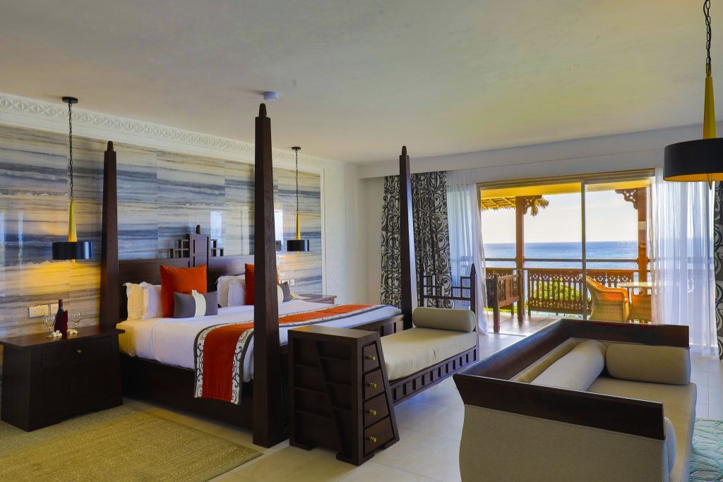 Zimmer der ocean-front-suite mit Himmelbett aus dunkelbraunem Holz, blau-grauen Wandpaneelen und Meerblick vom Balkon
